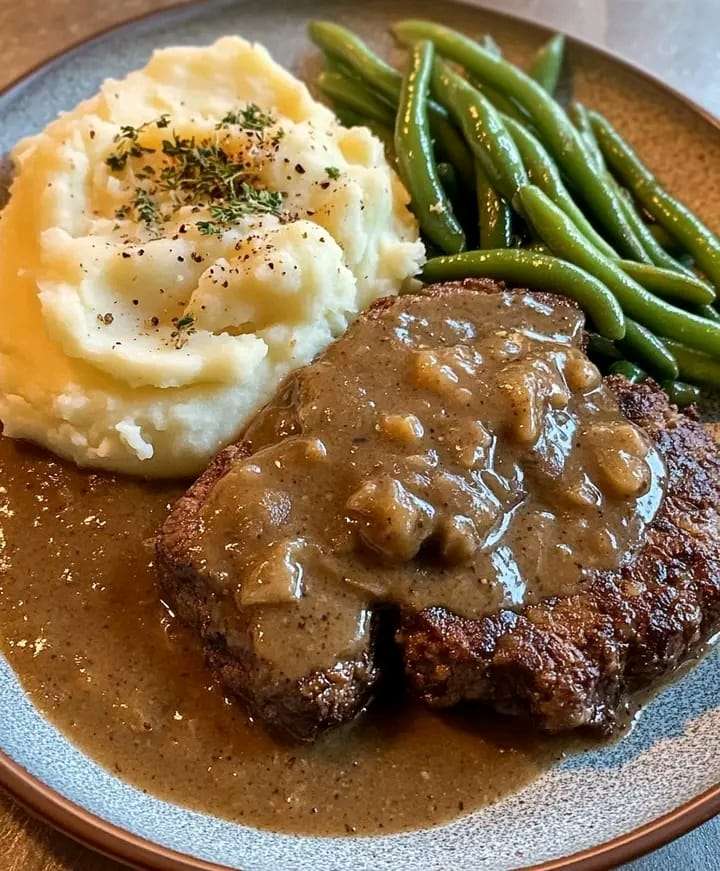 A plate with beef cube steak smothered in brown gravy, creamy mashed potatoes, and sautéed green beans.