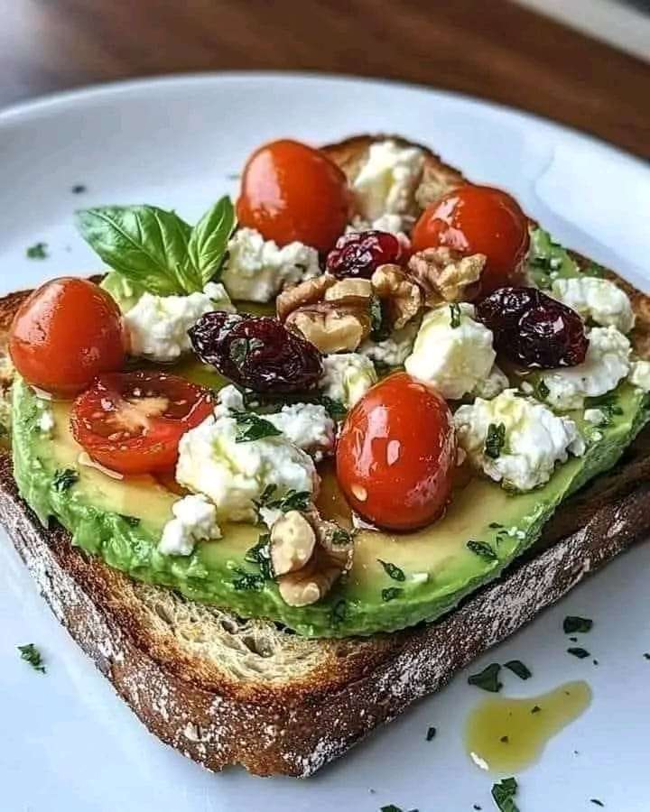 Whole-grain toast topped with mashed avocado, crumbled feta, cherry tomatoes, cranberries, and walnuts, garnished with fresh parsley.