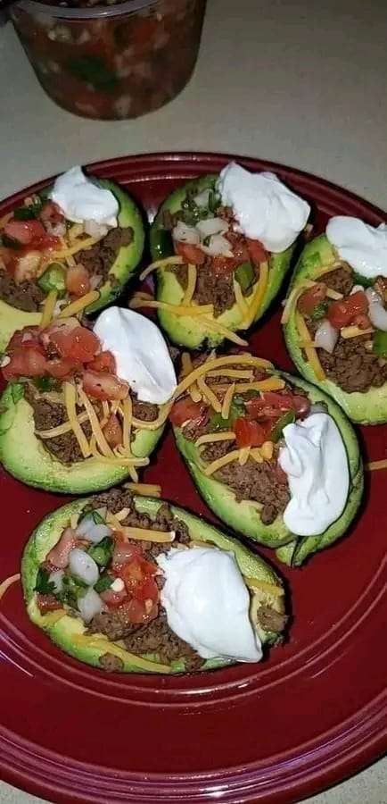 A halved avocado stuffed with seasoned beef, topped with salsa, sour cream, cheese, and lettuce, served on a white plate.