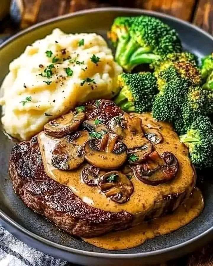 A juicy steak topped with creamy mushroom sauce, served with mashed potatoes and steamed broccoli on a plate.