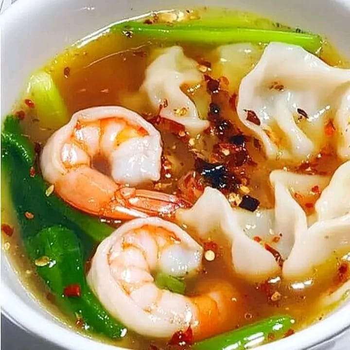 A steaming bowl of wonton soup filled with shrimp wontons, bok choy, and green onion, served in a white bowl.