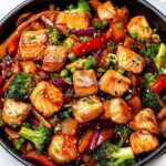 A colorful skillet of salmon stir fry with broccoli, carrots, and bell peppers, garnished with sesame seeds and green onions.