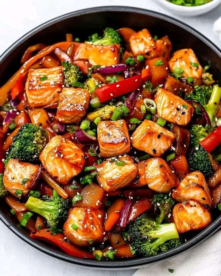 A colorful skillet of salmon stir fry with broccoli, carrots, and bell peppers, garnished with sesame seeds and green onions.