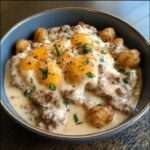 A hearty Tater Tot Breakfast Bowl topped with sausage gravy, scrambled eggs, and garnished with green onions and cheddar cheese.