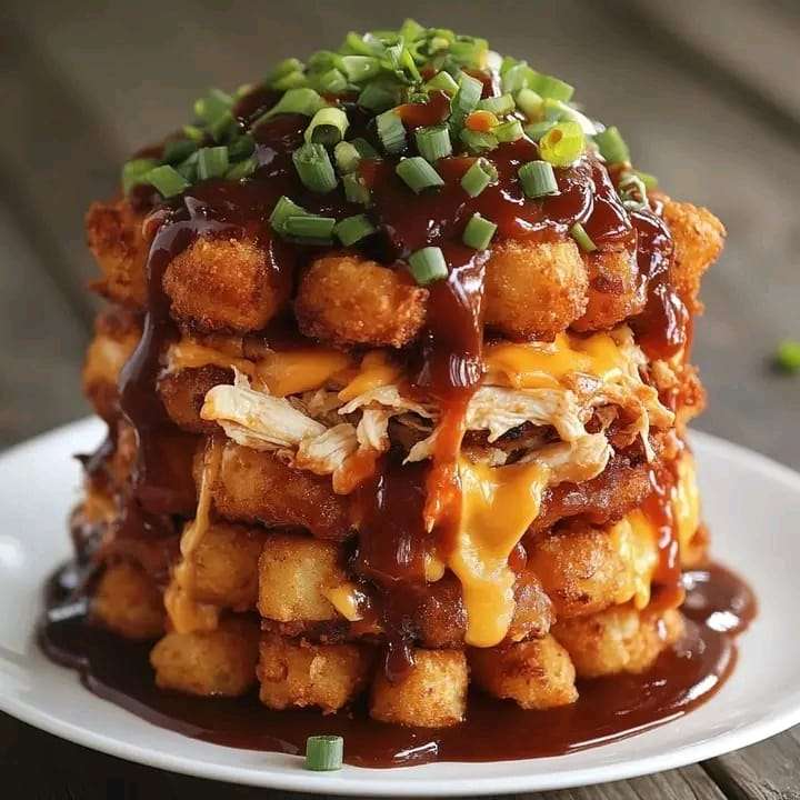 A stack of crispy tater tots layered with BBQ chicken, melted cheese, and a drizzle of BBQ sauce, garnished with green onions.