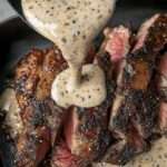 Sliced Peppercorn Crusted Steak drizzled with creamy peppercorn sauce, served alongside roasted potatoes and steamed broccoli.