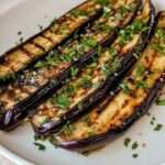 Grilled eggplant slices garnished with Parmesan and parsley, served on a white plate with a drizzle of balsamic glaze.