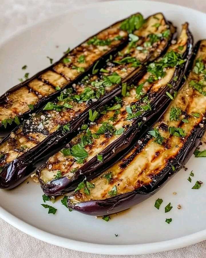 Grilled eggplant slices garnished with Parmesan and parsley, served on a white plate with a drizzle of balsamic glaze.