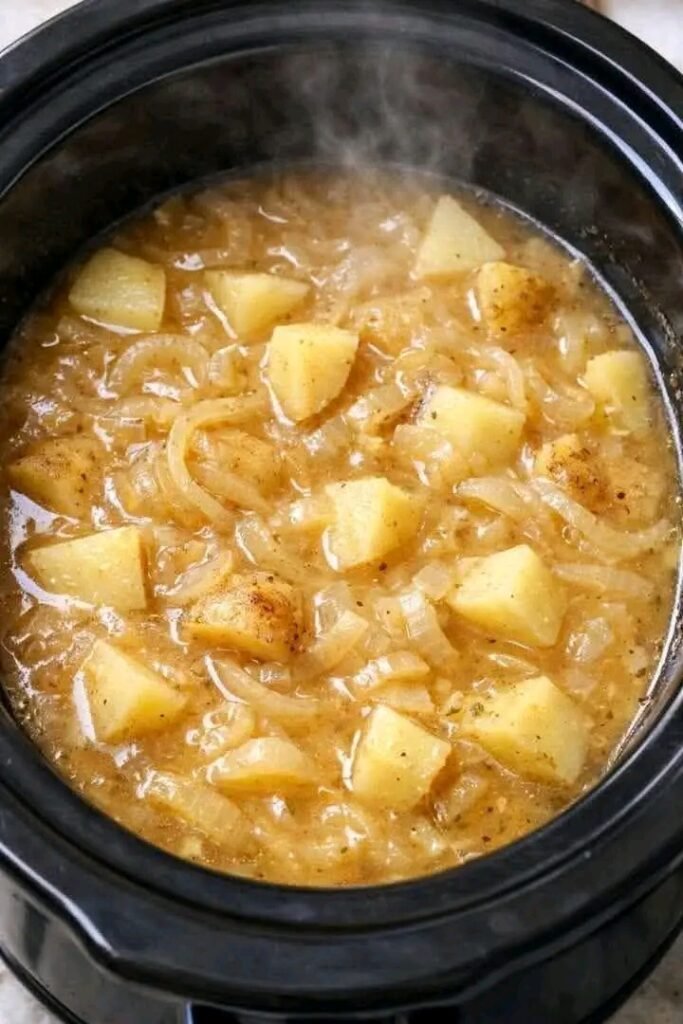 Steaming hot potato and onion stew simmering in a black slow cooker, featuring tender cubed potatoes and translucent onions in a savory golden broth.