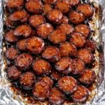 A foil-lined baking sheet piled with golden-brown, caramelized sliced sausage bites glazed in a sticky honey garlic sauce.