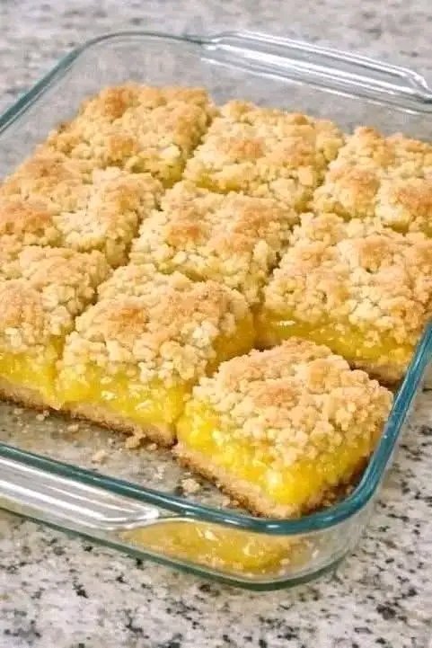 A glass baking dish filled with golden-brown lemon crumble bars, cut into squares, showcasing a vibrant yellow lemon filling and a textured crumb topping.
