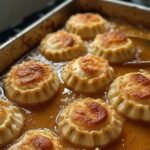 Jumbo cheese ravioli arranged neatly in a metal baking pan, simmered in a rich golden-brown savory sauce with crispy, toasted tops.
