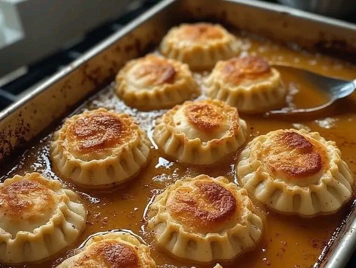 Jumbo cheese ravioli arranged neatly in a metal baking pan, simmered in a rich golden-brown savory sauce with crispy, toasted tops.