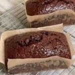 Two freshly baked, moist chocolate loaf cakes cooling on a wire rack, lined with brown parchment paper, showcasing a rich dark chocolate color and a glossy top crust.