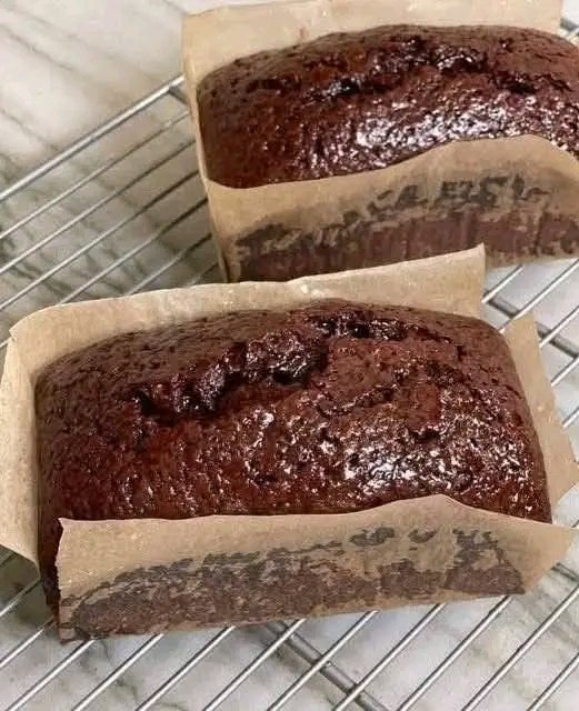 Two freshly baked, moist chocolate loaf cakes cooling on a wire rack, lined with brown parchment paper, showcasing a rich dark chocolate color and a glossy top crust.