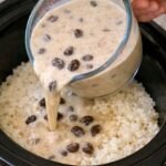 Creamy milk mixture with raisins and cinnamon being poured from a glass measuring cup over a bed of cooked white rice inside a black slow cooker.