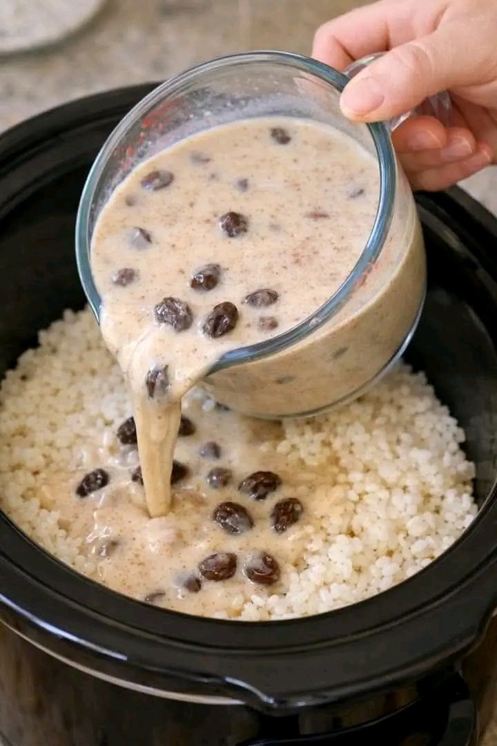 Creamy milk mixture with raisins and cinnamon being poured from a glass measuring cup over a bed of cooked white rice inside a black slow cooker.