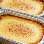 Two foil loaf pans filled with golden-brown baked milk pudding, showing a glossy custard surface dusted with ground cinnamon and caramelized, crispy edges.