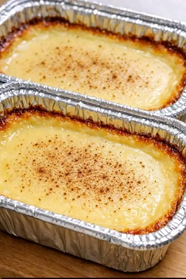 Two foil loaf pans filled with golden-brown baked milk pudding, showing a glossy custard surface dusted with ground cinnamon and caramelized, crispy edges.
